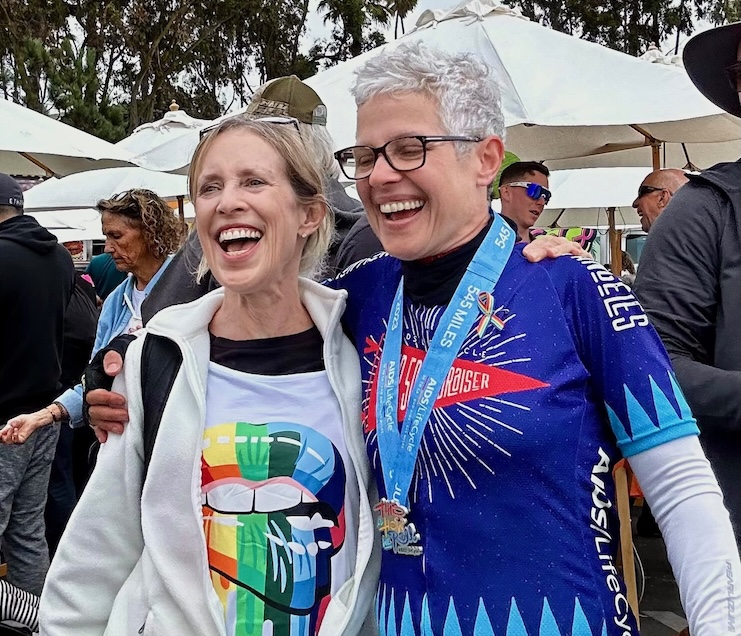 Eliza and Maggie laughing together at the 2023 AIDS/LifeCycle finish line in Santa Monica, CA, Maggie wearing her finisher medal