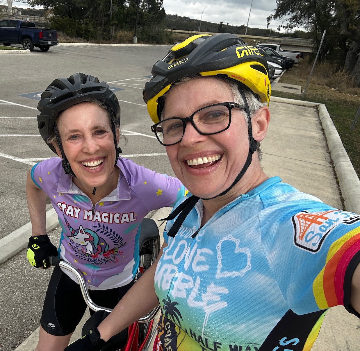 Eliza wearing a purple 'Stay Magical' unicorn cycling jersey and Maggie in a blue jersey, both grinning on their bikes